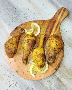Golden grilled chicken drumsticks with lemon slices on a wooden serving board, seasoned with herbs.