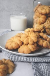 Golden fried flowers-shaped cookies on a white plate with a glass of milk and a jar of cookies in the background.