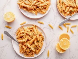 Creamy tomato pasta with chopped herbs on white plates with lemon wedges on marble surface.
