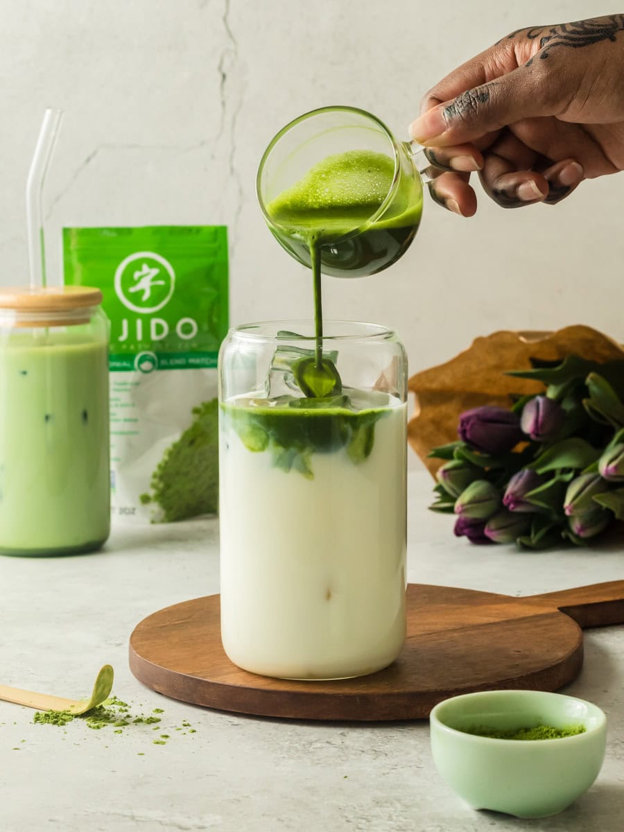 Bright green matcha being poured into a glass of milk with a matching green matcha powder jar in the background.