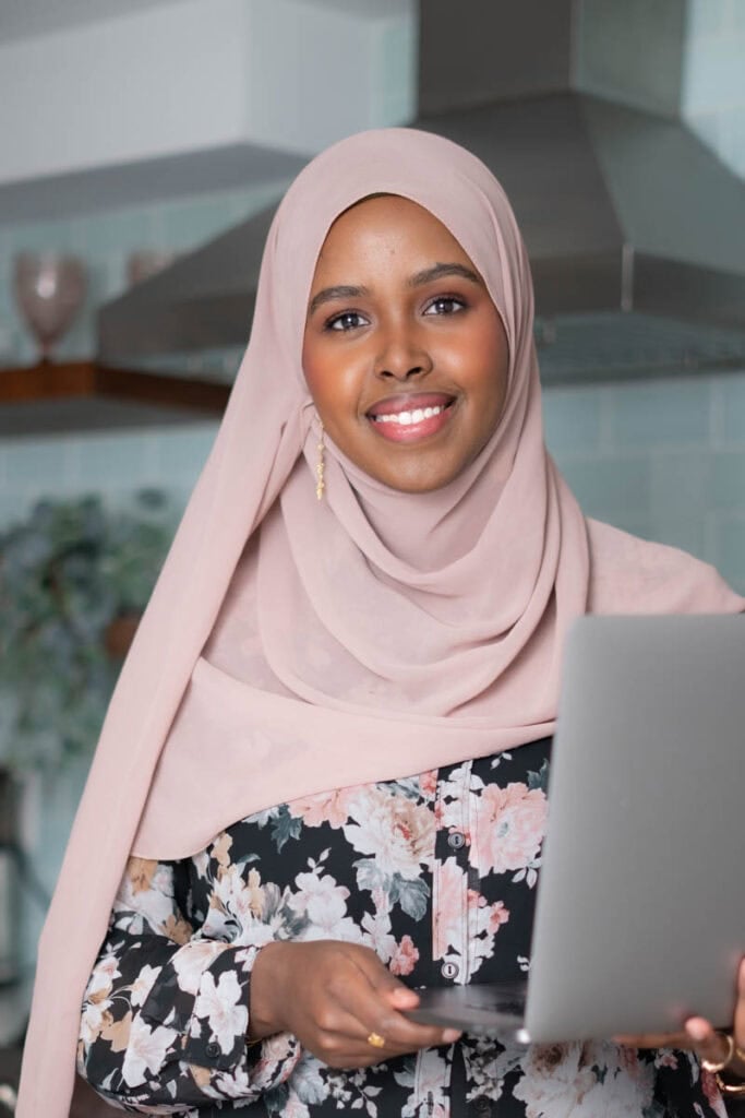 Smiling woman in hijab using a laptop in a modern kitchen.