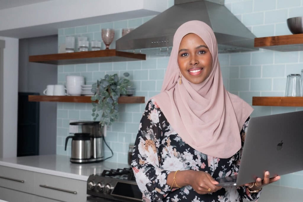 Bright smiling woman in hijab holding a laptop in a modern kitchen with pastel tiles.