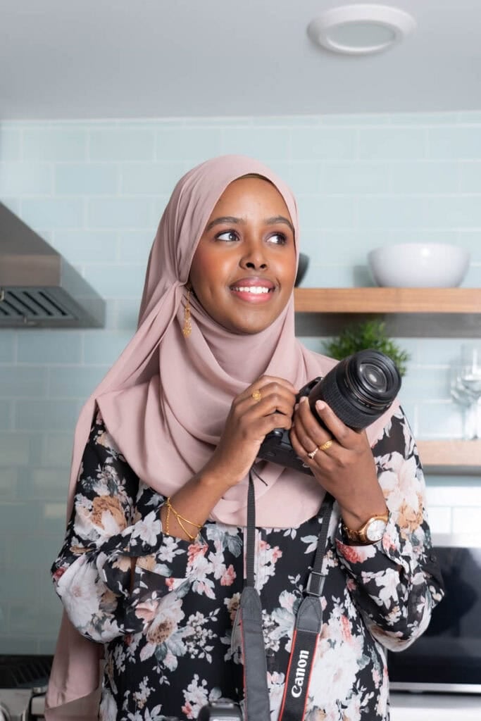 Female photographer wearing a pink hijab and floral dress, holding a camera in a modern kitchen.