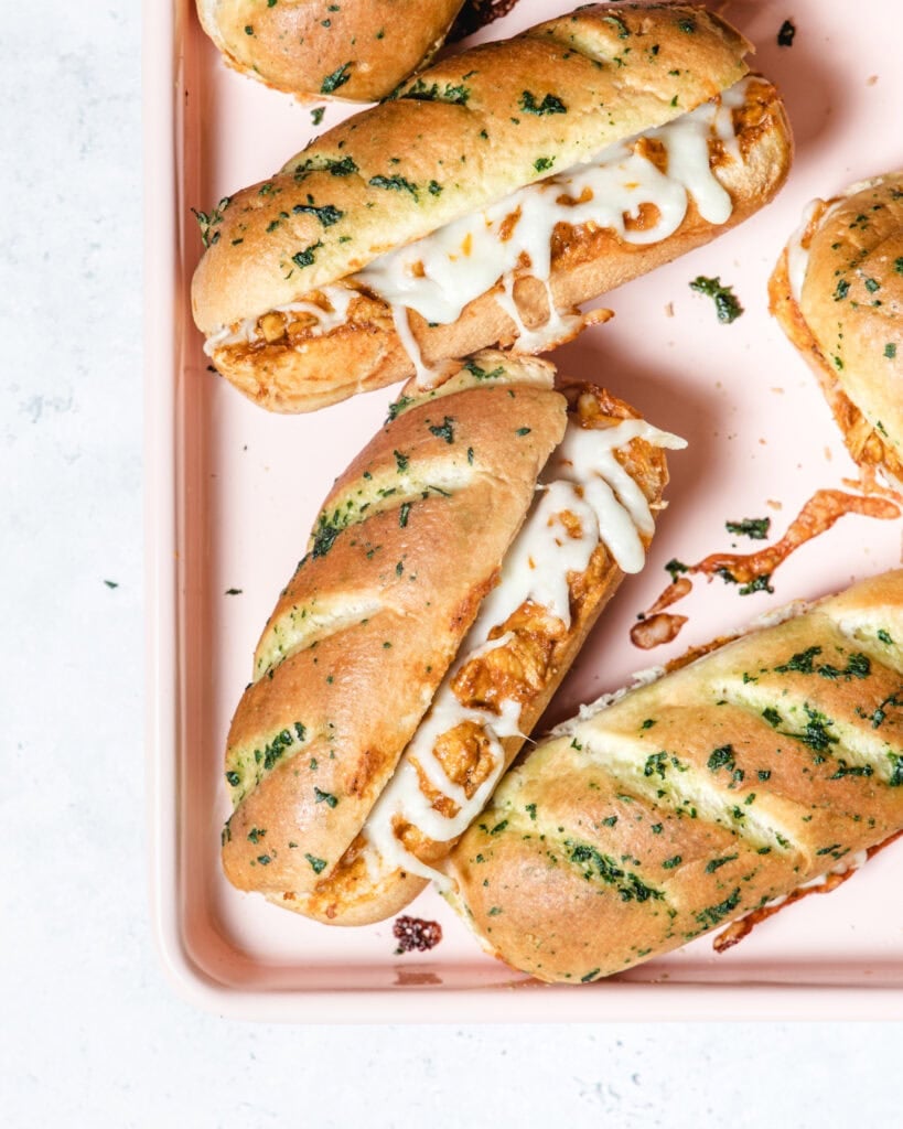 Three baguette sandwiches filled with melted cheese and herbs on a pink tray.