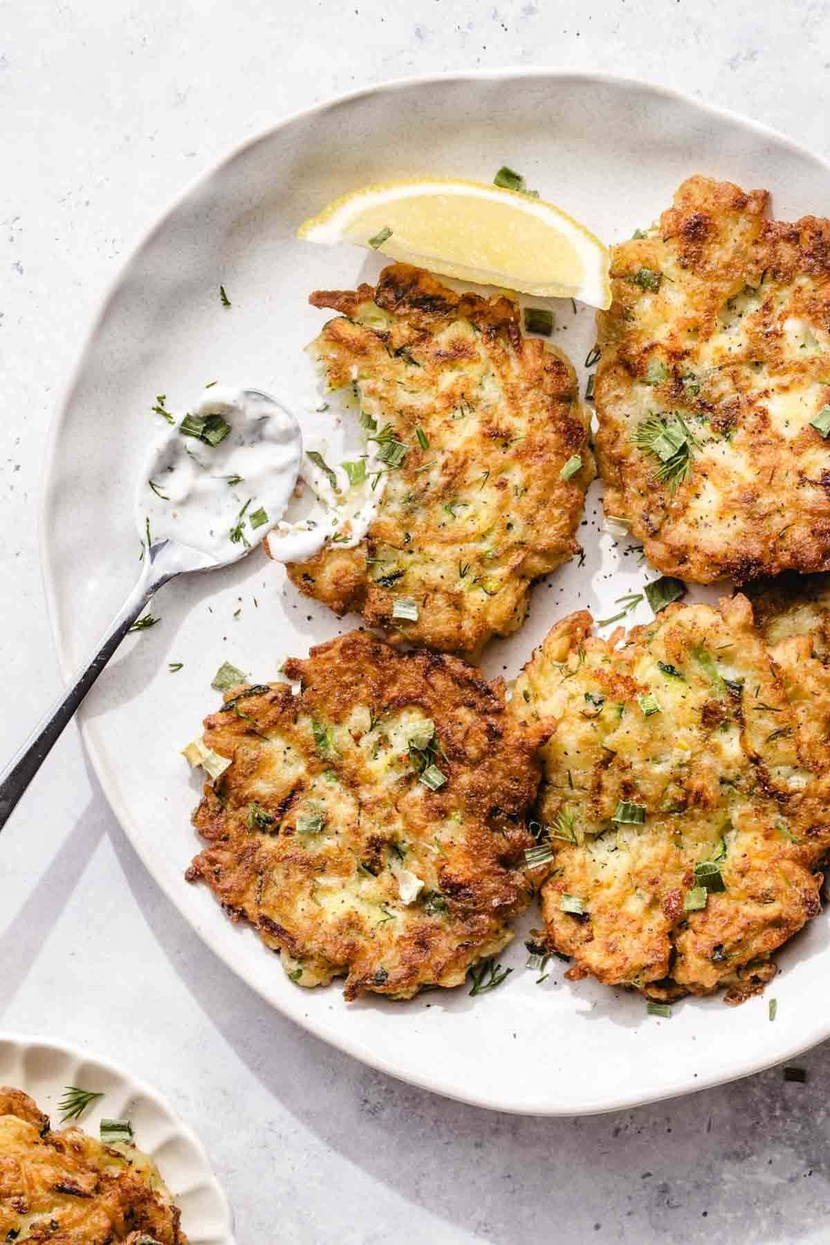 Four golden-brown fish fillets garnished with chopped herbs and a lemon wedge on a white plate.