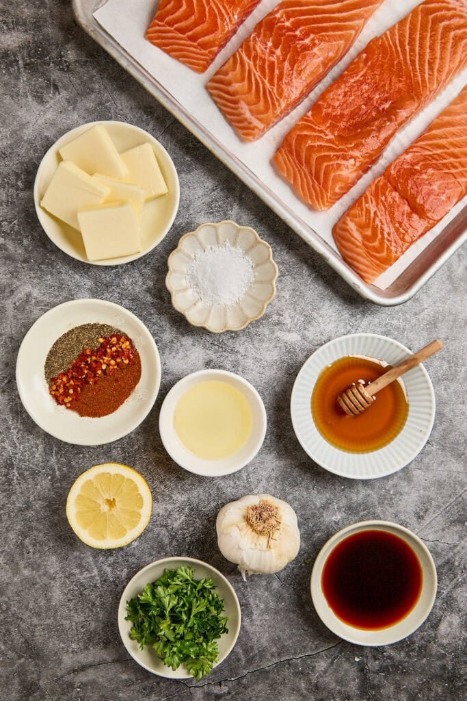 Fresh salmon fillets with spices, lemon, garlic, honey, and herbs on a gray surface.